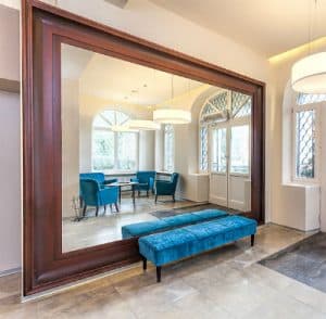 Huge, beautiful, custom mirror by Glass King in a luxury home entrance way, with a brown wooden frame and blue velvet bench in front.