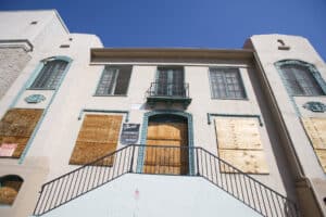 A beige two-story residential building with boarded-up windows and doors, featuring a small balcony and blue trim under a clear sky.