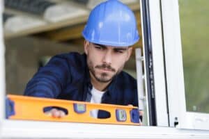 A construction worker in a blue hard hat uses a spirit level to check the alignment of a patio door frame.