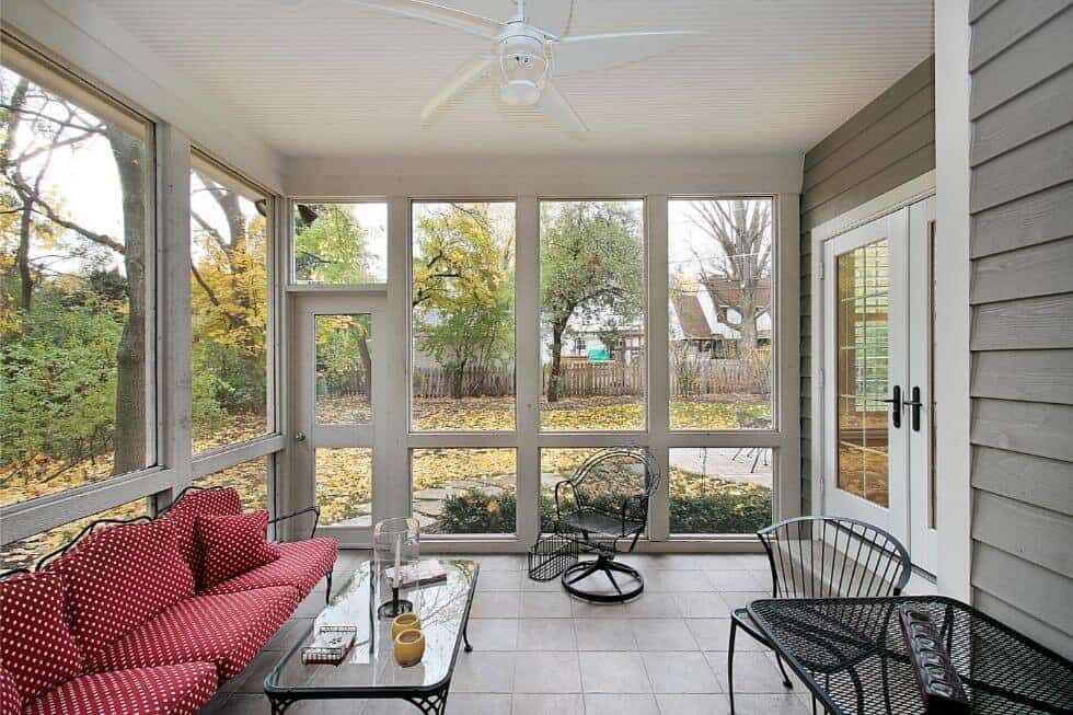 A cozy screened-in porch with bug screens, a red sofa, two black chairs, and a glass coffee table, overlooking a yard through large windows.