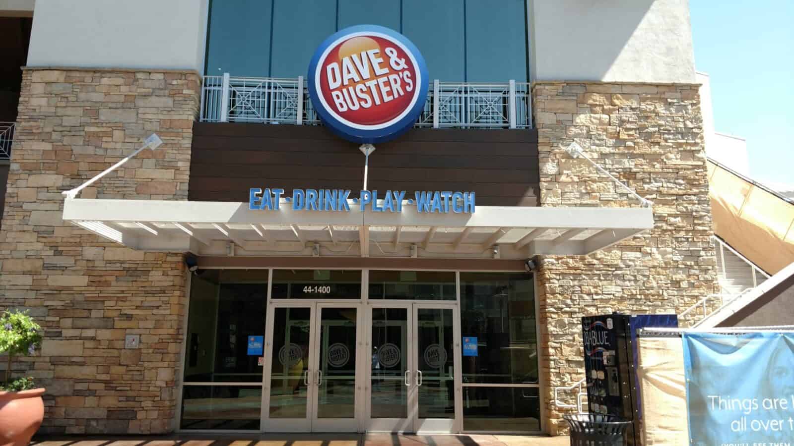 Exterior of a Dave & Buster's commercial entertainment venue with prominent "eat, drink, play, watch" sign and logo above the entrance doors.