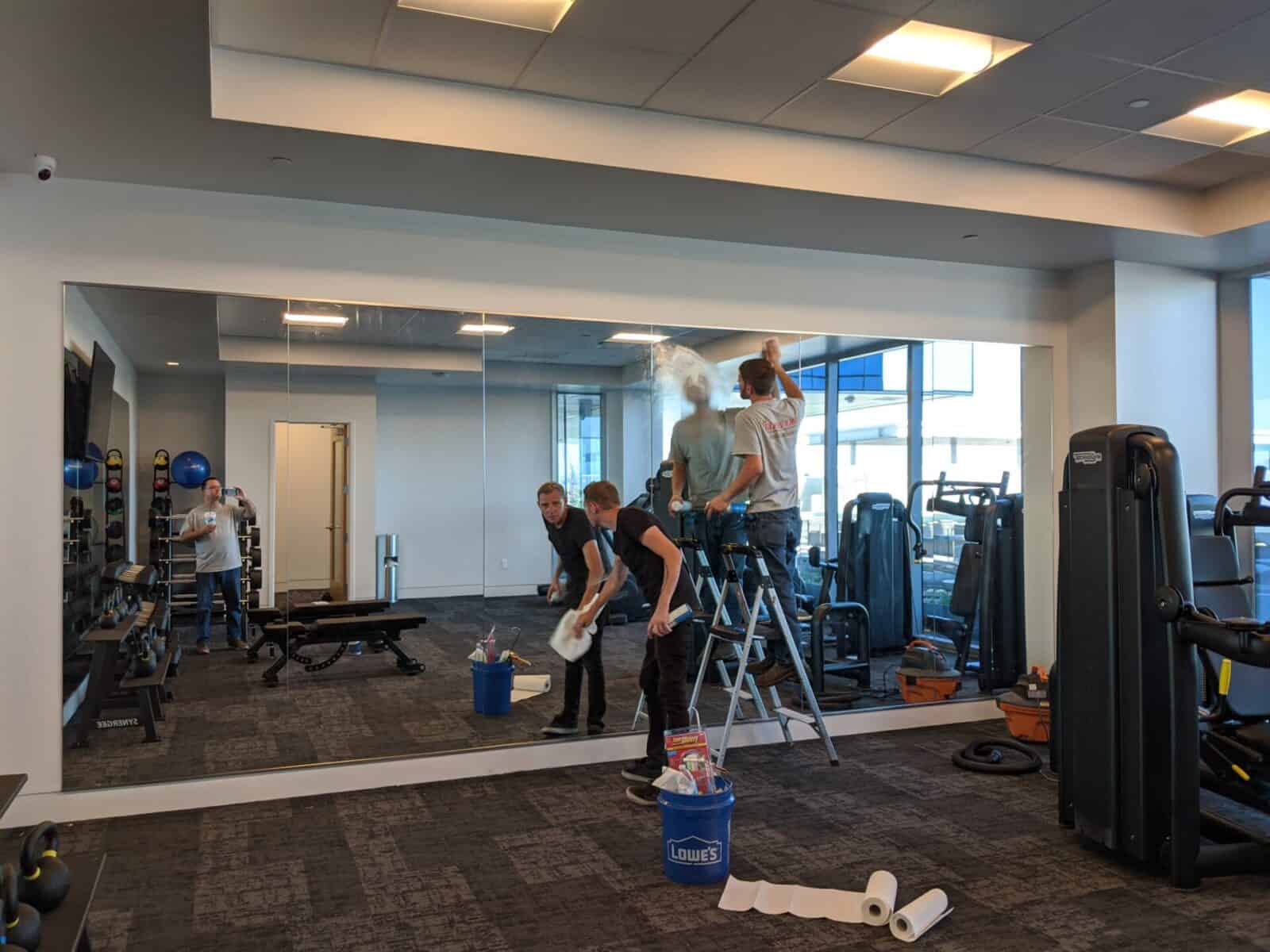 Workers install lighting in a gym with custom home gym mirrors and exercise equipment, as a man cleans the floor.