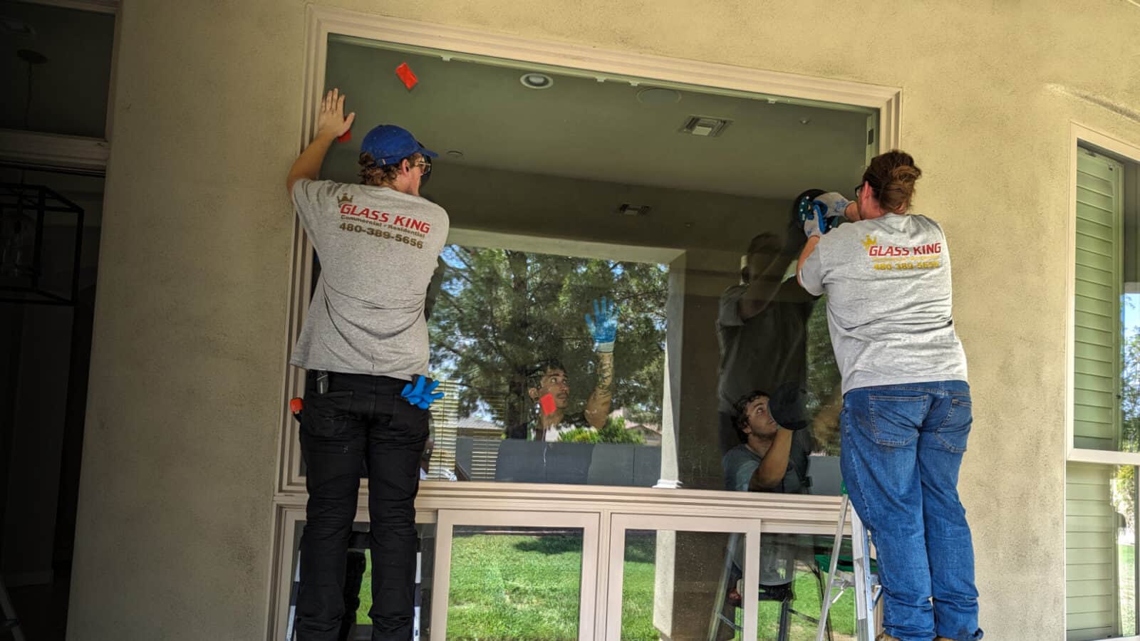Two workers perform a home window repair on a large exterior window; one secures the top while the other assists from the side.